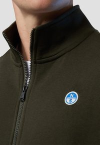 Giacca in pile verde scuro con zip e colletto alto, dotata di un logo circolare blu sul petto e hardware della cerniera visibile.