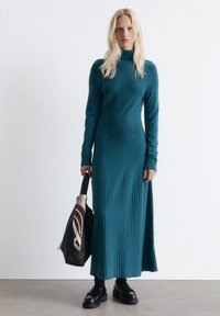 Eine Frau mit langen blonden Haaren, die ein türkisfarbenes, geripptes Strick-Turtleneck-Kleid trägt, schwarze Mokkasins anhat und eine schwarze Handtasche mit einem gemusterten Schal hält.