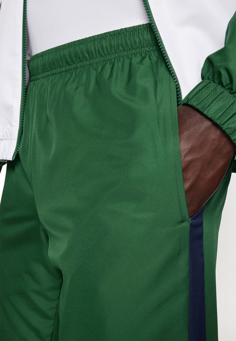 Pantaloni sportivi verdi con vita elastica, tasche laterali e finitura texturizzata. Presentano strisce laterali navy per un contrasto.