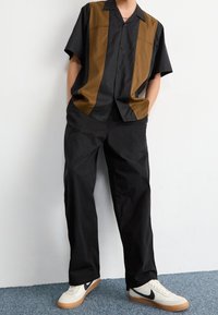 Koszula z krótkim rękawem w czarnym i brązowym kolorze, z zapinaniem na guziki i dwukolorowym wzorem; połączona z luźnymi czarnymi spodniami i białymi trampkami.