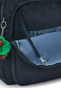 Mörkblå ryggsäck med en slät tygfinish, med en ficka med dragkedja och en grön fluffig nyckelring fäst. Blå inre ficka synlig.
