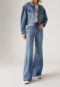 Gekürzte blaue Jeansjacke mit Knopfverschluss und Brusttaschen, kombiniert mit hochtaillierten, ausgestellten blauen Jeans und weißen Turnschuhen.