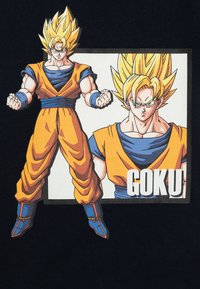 T-shirt orange et bleu mettant en avant Goku, un personnage d'anime aux cheveux jaunes, avec des muscles bien définis, prenant une pose, accompagné d'un texte imprimé en dessous.