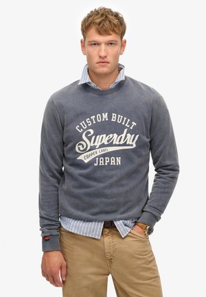 Grijze sweatshirt met witte "Superdry" logo, gedragen over een lichtblauwe gestreepte blouse. Bruine broek en een polshorloge zijn ook zichtbaar.