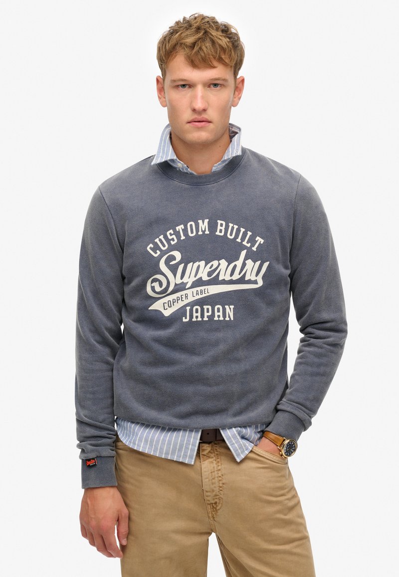 Sudadera gris con el logo blanco de "Superdry", llevada sobre una camisa de cuello a rayas en azul claro. También son visibles pantalones beige y un reloj de pulsera.