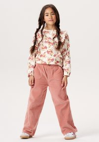 Długo-sleeve top w kwiatowy wzór z falbanami, połączony z luźnymi, różowymi spodniami z korduroju. Modelka nosi białe trampki z beżowymi podeszwami.