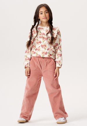 Haut à manches longues à motifs floraux avec des volants associé à un pantalon en velours côtelé rose ample. Le modèle porte des baskets blanches avec des semelles beiges.