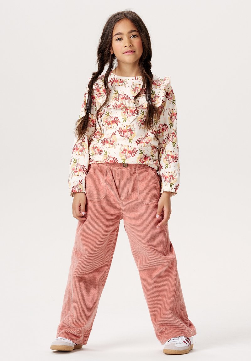 Długo-sleeve top w kwiatowy wzór z falbanami, połączony z luźnymi, różowymi spodniami z korduroju. Modelka nosi białe trampki z beżowymi podeszwami.