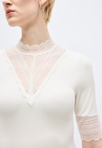 Ξανθό άτομο με λευκό τοπ με περίτεχνο λευκό γιακά από δαντέλα, V-σχήμα πάνελ δαντέλας στο στήθος και δαντελένιο τελείωμα στα κοντά μανίκια. Ορατό ασημένιο κρίκο σκουλαρίκι.