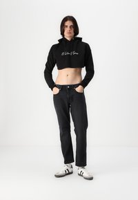 Cropped black hoodie with logo, paired with dark jeans and white sneakers featuring black stripes. Model stands against a plain white background.