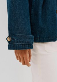 Rękaw ciemnoniebieskiej dżinsowej kurtki z detalem mankietu, charakteryzujący się zaokrąglonym kształtem i pojedynczym okrągłym guzikiem. Ręka umiejscowiona poniżej rękawa.