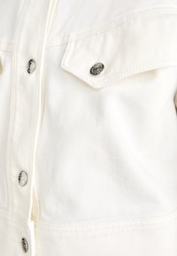White denim jacket featuring silver buttons, two chest pockets, and a textured fabric. Detail stitching visible along seams.