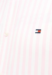 Ροζ ανοιχτό και λευκό πουκάμισο με κάθετες ρίγες και ένα μικρό κεντητό ορθογώνιο λογότυπο σε ναυτικό μπλε, λευκό και κόκκινο κοντά στα κουμπιά.