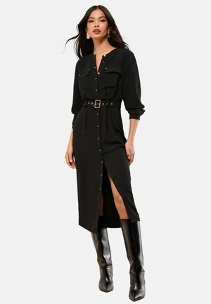 Schwarzes Hemdkleid mit langen Ärmeln, Knopfleiste, zwei Brusttaschen, Gürtel mit goldener Schnalle und seitlichem Schlitz, kombiniert mit glänzenden kniehohen Stiefeln.