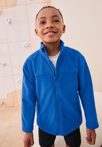 Blå fleecejakke med lynlås, høj krave, sidelommer og flade sømme. Bruges over en hvid skjorte, kombineret med sorte shorts.