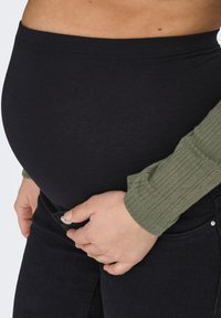 Melns, elastīgs materiāls grūtnieču jostasvietai pār apaļu vēderu, saskaņots ar teksturētu, garām piedurknēm olīvzaļu topu un tumšām džinsām.