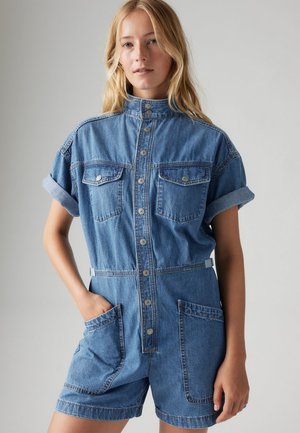 Tuta in denim con maniche corte, colletto, bottoni frontali, due tasche sul petto e due tasche laterali, in una tonalità di blu chiaro.