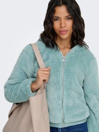 Jeune femme portant une veste zippée bleu clair et duveteuse, avec un grand sac fourre-tout beige sur l'épaule.