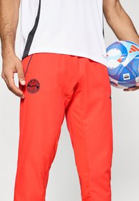 Czerwone spodnie sportowe o gładkiej fakturze, z okrągłym logo FC Bayern München na lewej udzie. Połączone z białym sportowym topem.