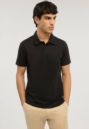 Mladý muž s tmavými kudrnatými vlasy, oblečený v černém polo tričku s krátkým rukávem a béžových kalhotách, stojící s rukama v kapsách, hledící doprava.