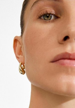 Ohrringe in Goldoptik mit wellenförmigem Design, polierter Oberfläche, kreisförmiger Form und dezentem Muster. Die Befestigungen sitzen nah am Ohr.