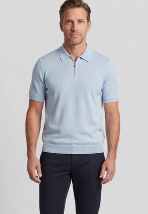 Man draagt een lichtblauw poloshirt met korte mouwen en rits, en donkere broek, staand tegen een effen lichtgrijze achtergrond.