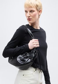 Άτομο με κοντά ξανθά μαλλιά, φορώντας μαύρο μακρυμάνικο πουκάμισο και λευκό παντελόνι, κρατώντας μια μαύρη δερμάτινη τσάντα ώμου σε λευκό φόντο.