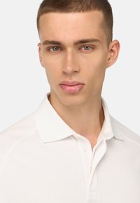 Bílá polo košile s límcem na knoflík, krátkými rukávy a texturovanou tkaninou. Střih vypadá jako na míru, zvýrazňuje ramena.