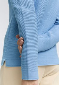Pull bleu clair avec un tissu à côtes texturé, des manches ajustées et un design court. Le corps présente un détail d'ourlet fendu.