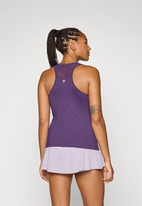 En kvinne med flettet hår iført en lilla top og en lavendel skort står med ryggen til kameraet mot en ensfarget bakgrunn.