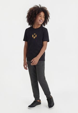 Mosolygós gyerek göndör hajjal, fekete grafikás pólóban, sötétszürke melegítőnadrágban és fekete sportcipőben, fehér háttér előtt állva.