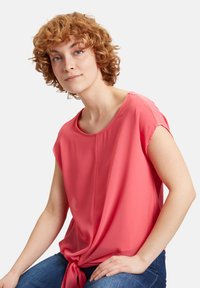 Top corto rosa a maniche corte con scollatura rotonda, caratterizzato da un dettaglio annodato in vita. Il tessuto appare leggero e leggermente lucido.