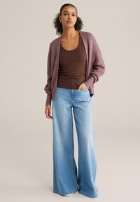 Cardigan mov deschis peste un top maro, asortat cu blugi albastri cu picioare largi. Cardiganul este realizat dintr-un material tricotat texturat și are un croi lejer.