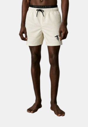 Uomo che indossa pantaloncini da bagno beige con coulisse nera e logo nero sulla gamba sinistra, in piedi a piedi nudi su sfondo semplice.