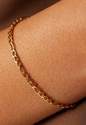 Gouden kettingarmband met ovale schakels, reflecterend oppervlak en een gladde textuur, gedragen om een gebruinde pols tegen een neutrale achtergrond.