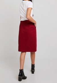 Falda lápiz burdeos de algodón, hasta la rodilla con bolsillos laterales, combinada con una blusa blanca ajustada y botines negros. Textura suave.