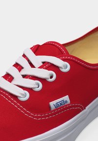 Κόκκινο αθλητικό παπούτσι Vans με λευκά κορδόνια, λευκή ραφή και λευκή λαστιχένια σόλα, εμφανίζοντας την ετικέτα του λογότυπου στο πλάι κοντά στα κορδόνια.