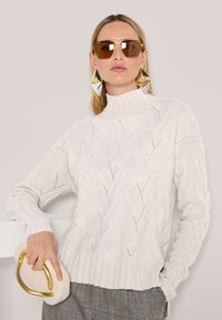 Eine Frau trägt einen weißen, strukturierten Rollkragenpullover, große geometrische goldene Ohrringe, braune Sonnenbrille und hält eine cremefarbene Clutch mit einem goldenen Griff.