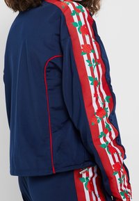 Marinblå idrottsjacka med röd blommig broderi på vitrandiga ärmar, texturerat material och röda detaljer längs sömmar och dragkedjor.