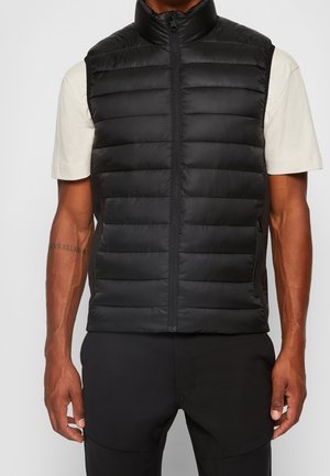 Mand iført sort quiltet ærmeløs vest lynet op over lysebeige kortærmet skjorte, står med armene afslappet ned langs siden.