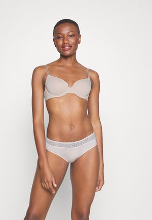 Femme portant un soutien-gorge beige et des sous-vêtements assortis avec des bordures en dentelle, se tenant devant un fond blanc uni, souriant doucement.