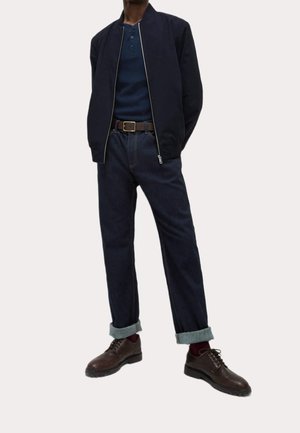 Mand iført en navy bomberjakke, mørkeblå henley-shirt, opkrogede mørke jeans, brun bælte, vinrøde sokker og brune lædersko til fest.