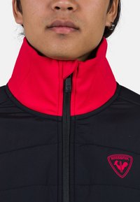 Černá bundu s péřovým designem a vysokým červeným límcem, předním zipem a červenou nášivkou loga Rossignol na hrudi. Hladká textura, moderní design.