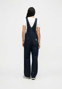 Γυναίκα με μακριά μαύρα μαλλιά, φοράει σκούρα τζιν ολόσωμη φόρμα με φαρδιά πόδια και λευκό κοντομάνικο πουκάμισο, στέκεται με την πλάτη προς τα εμπρός σε απλό φόντο.