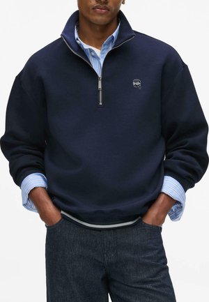 Man draagt een marineblauwe quarter-zip trui over een blauw-wit gestreepte blouse met kraag en donkere spijkerbroek, handen in de zakken.