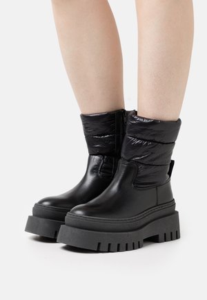 Botas negras con un upper acolchado y brillante, y una suela de goma gruesa y estriada. El diseño no tiene cordones y cuenta con un zipper lateral para facilitar su uso.