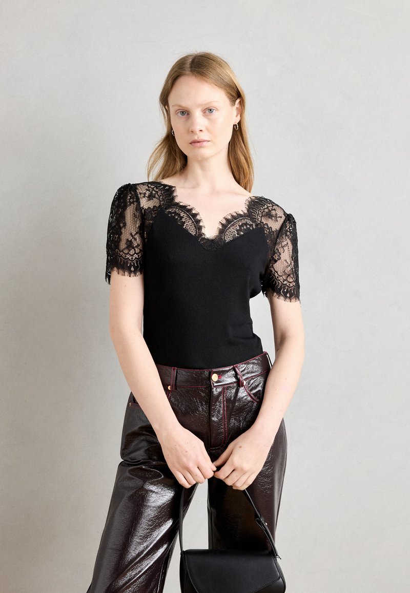 Haut noir avec des manches courtes en dentelle, présentant un col en V et un corsage ajusté. Assorti à un pantalon bordeaux brillant et un petit sac à main noir.