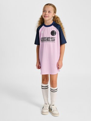 Ung pige med krøllet hår i en pink og marineblå sportsjersey kjole, hvide knæstrømper med sorte striber og sneakers, stående og kigger op.