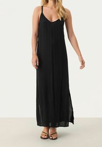 Schwarzes Maxikleid mit dünnen Trägern, V-Ausschnitt und seitlichen Schlitzen. Aus leichtem Stoff für ein fließendes Design. Flache schwarze Sandalen runden den Look ab.