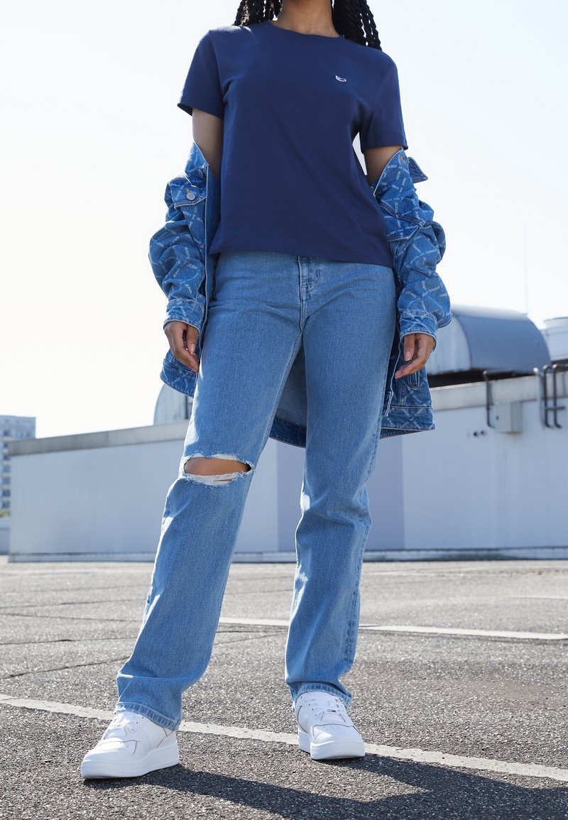 Persona con camiseta azul marino, vaqueros claros desgarrados, zapatillas blancas y una chaqueta vaquera sobre los hombros, de pie al aire libre sobre asfalto.